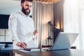 Young bearded smiling businessman in white shirt is standing near desk in front of laptop, holding in his hand documents Royalty Free Stock Photo