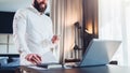 Young bearded smiling businessman in white shirt is standing near desk in front of laptop, holding in his hand documents Royalty Free Stock Photo