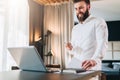 Young bearded smiling businessman in white shirt is standing near desk in front of laptop, holding in his hand documents Royalty Free Stock Photo