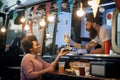 Young beardy caucasian employee in fast food laughing, adding a mustard in a sandwich to a female afro-american customer