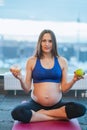 Young beauteous pregnant woman in blue sportswear sit on pink mat choosing between apple and doughnut at gym. Vertical.