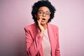 Young beautiful African American afro businesswoman with curly hair wearing pink jacket hand on mouth telling secret rumor, Royalty Free Stock Photo