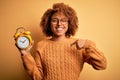 Young beautiful African American afro woman with curly hair holding vintage alarm clock with surprise face pointing finger to Royalty Free Stock Photo