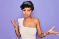 Young beautiful african american afro woman wearing tiara crown over purple background clueless and confused expression with arms Royalty Free Stock Photo