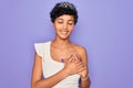 Young beautiful african american afro woman wearing tiara crown over purple background smiling with hands on chest with closed Royalty Free Stock Photo