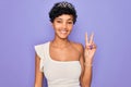 Young beautiful african american afro woman wearing tiara crown over purple background smiling with happy face winking at the Royalty Free Stock Photo