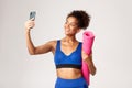 Young beautiful african-american fitness woman in sport outfit, taking selfie with yoga mat, taking photo on smartphone Royalty Free Stock Photo