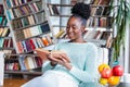 Young beautiful African American girl reading a book on the couch with the library bookshelves in the back. Beautiful woman on a Royalty Free Stock Photo