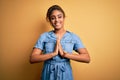 Young beautiful african american girl wearing denim dress standing over yellow background praying with hands together asking for Royalty Free Stock Photo