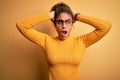 Young beautiful african american girl wearing sweater and glasses over yellow background Crazy and scared with hands on head, Royalty Free Stock Photo