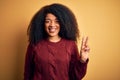 Young beautiful african american woman with afro hair standing over yellow isolated background smiling with happy face winking at Royalty Free Stock Photo