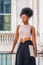 Young beautiful African American woman with afro hairstyle wearing sleeveless light color top, belt, black skirt, holding laptop Royalty Free Stock Photo