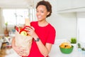 Young beautiful african american woman holding paper bag full of fresh healthy groceries at the kitchen Royalty Free Stock Photo