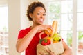 Young beautiful african american woman holding paper bag full of fresh healthy groceries and picking vegetables Royalty Free Stock Photo