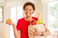 Young beautiful african american woman holding paper bag full of fresh healthy groceries and picking vegetables Royalty Free Stock Photo