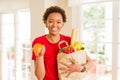 Young beautiful african american woman holding paper bag full of fresh healthy groceries and picking vegetables Royalty Free Stock Photo