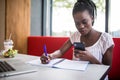 Young beautiful Afro-American woman using laptop and writing in notebook while studying in cafe Royalty Free Stock Photo