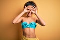 Young beautiful american woman on vacation wearing bikini over isolated yellow background doing ok gesture like binoculars Royalty Free Stock Photo