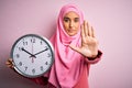 Young beautiful arab woman wearing pink muslim hijab doing countdown holding big clock with open hand doing stop sign with serious Royalty Free Stock Photo