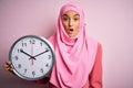 Young beautiful arab woman wearing pink muslim hijab doing countdown holding big clock scared in shock with a surprise face, Royalty Free Stock Photo