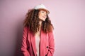 Young beautiful architect woman with curly hair wearing safety helmet over pink background looking away to side with smile on Royalty Free Stock Photo