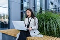 Young beautiful Asian business woman working in the air on a laptop, uses a headset for a video call, sitting near a modern office Royalty Free Stock Photo