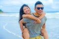 Young beautiful Asian Chinese couple with boyfriend carrying woman on her back and shoulders at the beach smiling