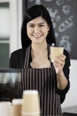 Young and beautiful Asian female barista wearing an apron standing in front of coffee machine. She holding offering paper coffee Royalty Free Stock Photo