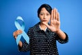 Young beautiful asian girl holding blue cancer ribbon symbol over isolated background with open hand doing stop sign with serious Royalty Free Stock Photo
