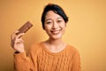 Young beautiful asian girl holding energy protein bar of cereal over isolated yellow background with a happy face standing and Royalty Free Stock Photo