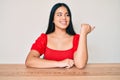 Young beautiful asian girl wearing casual clothes sitting on the table smiling with happy face looking and pointing to the side Royalty Free Stock Photo