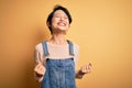 Young beautiful asian girl wearing casual denim overalls over isolated yellow background very happy and excited doing winner Royalty Free Stock Photo