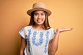 Young beautiful asian girl wearing casual t-shirt and hat standing over yellow background smiling cheerful presenting and pointing Royalty Free Stock Photo