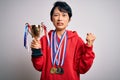 Young beautiful asian girl winner holding trophy wearing medals over white background annoyed and frustrated shouting with anger, Royalty Free Stock Photo