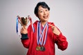 Young beautiful asian girl winner holding trophy wearing medals over white background very happy pointing with hand and finger Royalty Free Stock Photo