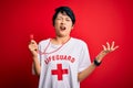 Young beautiful asian lifeguard girl wearing t-shirt with red cross using whistle crazy and mad shouting and yelling with