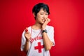 Young beautiful asian lifeguard girl wearing t-shirt with red cross using whistle looking stressed and nervous with hands on mouth