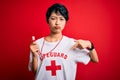 Young beautiful asian lifeguard girl wearing t-shirt with red cross using whistle Pointing down looking sad and upset, indicating