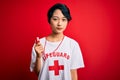 Young beautiful asian lifeguard girl wearing t-shirt with red cross using whistle Relaxed with serious expression on face