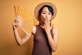 Young beautiful asian tourist girl on vacation wearing summer hat using hand fan cover mouth with hand shocked with shame for