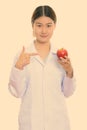 Young beautiful Asian woman doctor holding and pointing at red apple while looking at camera Royalty Free Stock Photo