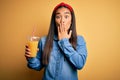 Young beautiful asian woman drinking healthy glass of orange juice over yellow background cover mouth with hand shocked with shame Royalty Free Stock Photo