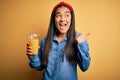 Young beautiful asian woman drinking healthy glass of orange juice over yellow background pointing and showing with thumb up to Royalty Free Stock Photo