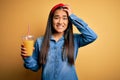 Young beautiful asian woman drinking healthy glass of orange juice over yellow background stressed with hand on head, shocked with Royalty Free Stock Photo