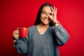 Young beautiful asian woman drinking mug of coffee standing over isolated red background with happy face smiling doing ok sign Royalty Free Stock Photo