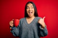 Young beautiful asian woman drinking mug of coffee standing over isolated red background pointing and showing with thumb up to the Royalty Free Stock Photo