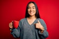 Young beautiful asian woman drinking mug of coffee standing over isolated red background with surprise face pointing finger to Royalty Free Stock Photo