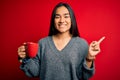 Young beautiful asian woman drinking mug of coffee standing over isolated red background very happy pointing with hand and finger Royalty Free Stock Photo