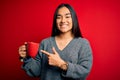 Young beautiful asian woman drinking mug of coffee standing over isolated red background very happy pointing with hand and finger Royalty Free Stock Photo