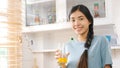 Young beautiful asian woman drinking orange juice, smiling and looking at camera while standing by window in kitchen background, Royalty Free Stock Photo